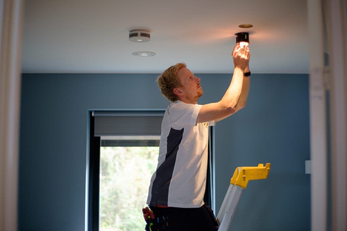 Røykvarsler i taket i en bolig mens elektrikeren monterer en downlight ved siden av